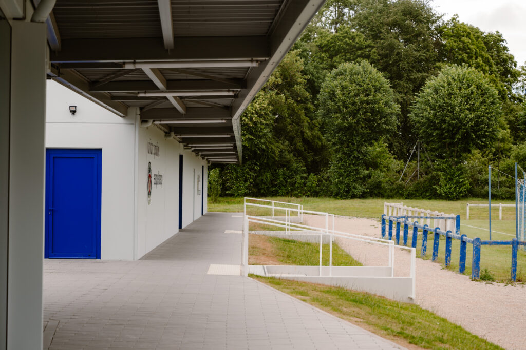 Moderne industriebouw met wit gevel en blauwe deuren naast sportveld omgeven door groen
